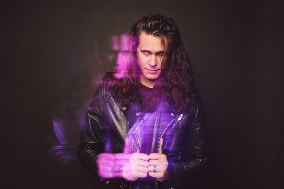 A person with long hair in a black leather jacket stands against a dark background. A purple and pink light effect creates a double exposure, showing a blurred duplicate of their face and upper body.