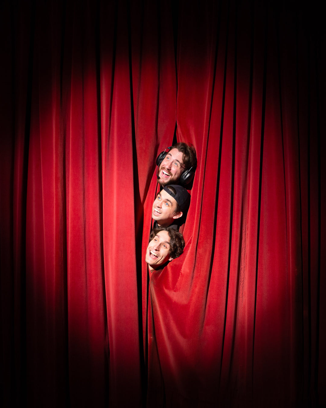 Three men poke their heads through an opening in tall red stage curtains, smiling and making playful expressions under dramatic theater lighting.