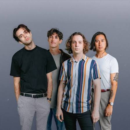 Four young men stand together against a plain gray background. They wear casual clothes, including t-shirts, jeans, and a striped shirt, and pose closely, looking at the camera with neutral expressions.