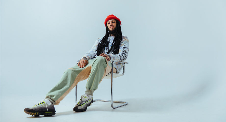 A person with long dreadlocks wearing a red beanie, patterned shirt, beige and mint green pants, and sneakers sits relaxed in a modern metal chair against a plain light background.