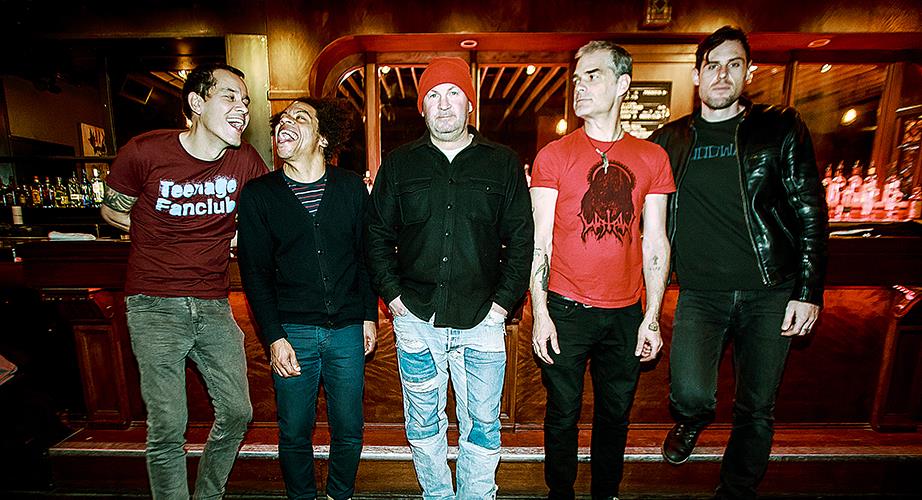 Five men stand in front of a bar; two are laughing while the others look serious. They wear casual clothes, including graphic t-shirts, jeans, and jackets, with warm lighting in the background.