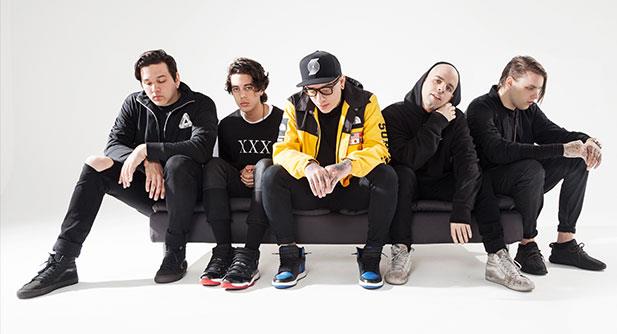 Five young men dressed in casual, dark clothing sit on a sofa against a plain white background, some looking down or away with relaxed and thoughtful expressions.