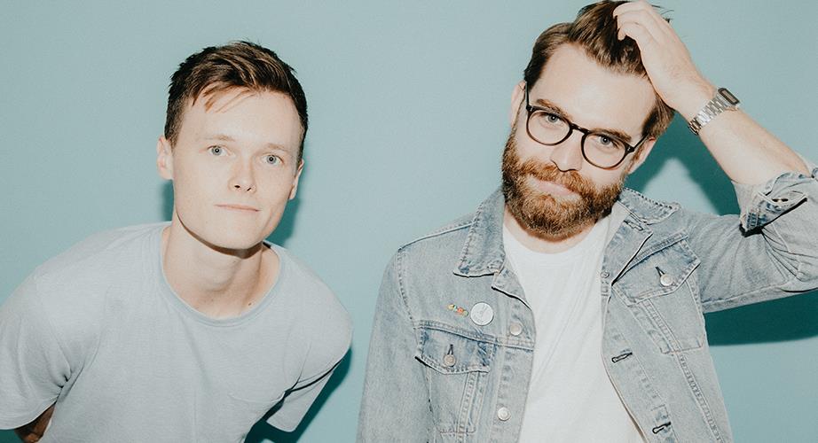 Two young men stand side by side against a light blue background. One has short hair and wears a light t-shirt. The other, with glasses and a beard, wears a denim jacket and touches his hair.