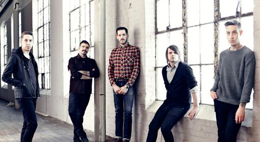 Five men stand and sit along large windows in an industrial-style room with white brick walls, wearing casual clothing and looking toward the camera.