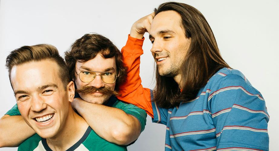 Three young men pose playfully; one with glasses and a mustache wraps his arms around a smiling friend, while the third, with long hair, smiles and interacts with the group.