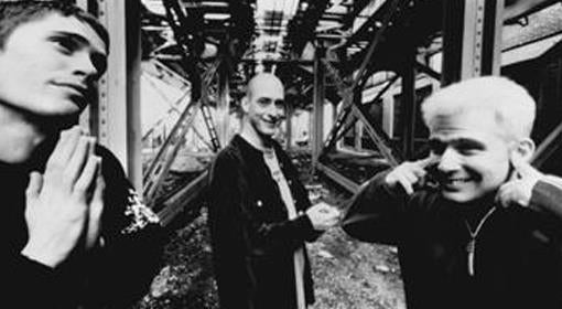 Three men stand under a metal structure; one poses with hands in a prayer gesture, one smiles at the camera, and the third plugs his ears and grins. The photo is in black and white.