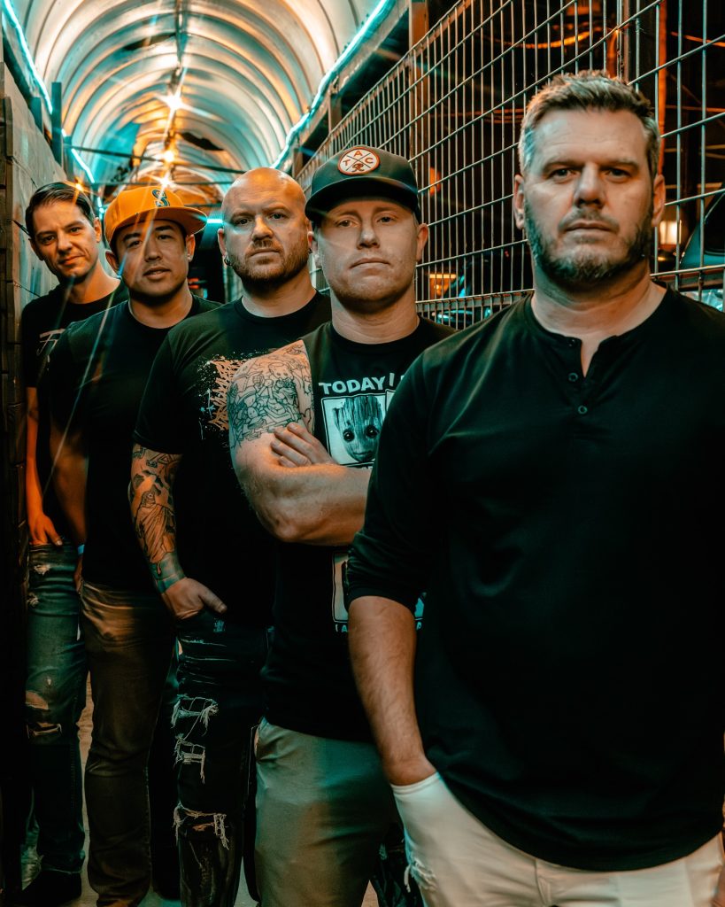 Five young men stand facing the camera, some with arms crossed, wearing casual clothes including t-shirts, hoodies, and jeans, against a plain, light-colored wall.