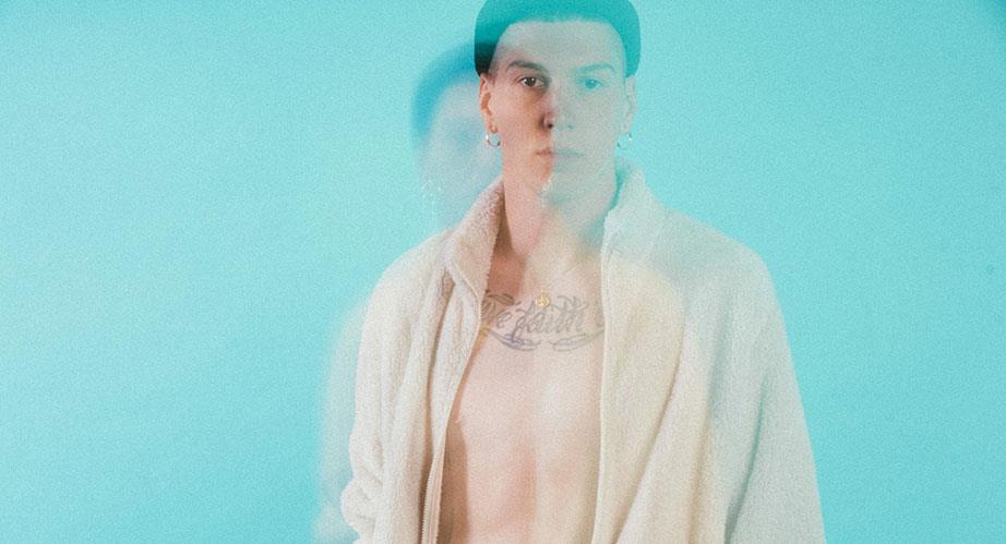 A young person with short dark hair, earrings, and a white robe stands against a blue background. The image is blurred, creating a double-exposure effect with a faint second outline.