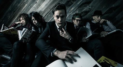 Five people sit closely together on a couch, dressed in dark clothing, reading magazines or books. The person in the center looks directly at the camera, reaching toward a book in front of them.