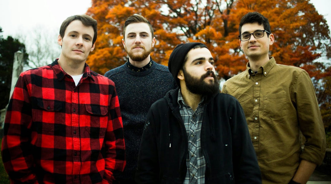 Four men stand outside in front of a tree with orange autumn leaves. They wear casual clothes, including a red plaid shirt, gray sweater, black jacket, and tan button-down shirt.