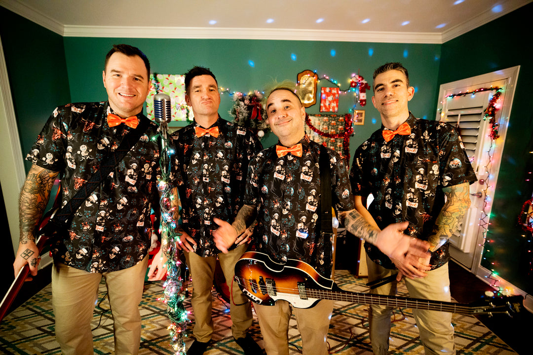Four men in matching Star Wars shirts and orange bow ties pose with musical instruments in a festive, colorfully decorated room with holiday lights and ornaments.