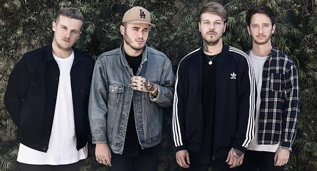 Four young men stand side by side outdoors in front of leafy greenery, dressed in casual streetwear including jackets, a denim shirt, and a baseball cap, looking directly at the camera.