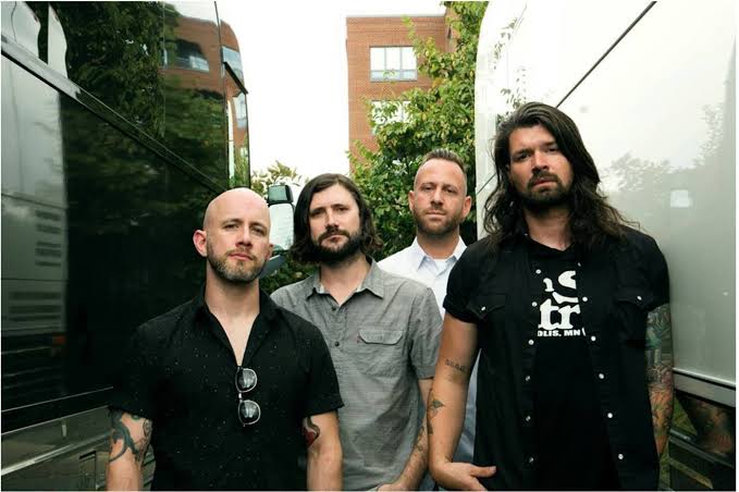 Four men with tattoos sit on a curb in front of green foliage, posing casually. They wear casual clothes, including jeans and T-shirts, with relaxed and friendly expressions.
