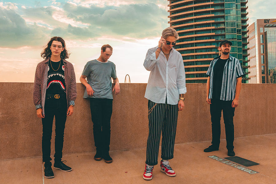 Four men stand on a rooftop, posing casually in trendy outfits, with modern buildings and a partly cloudy sky in the background.