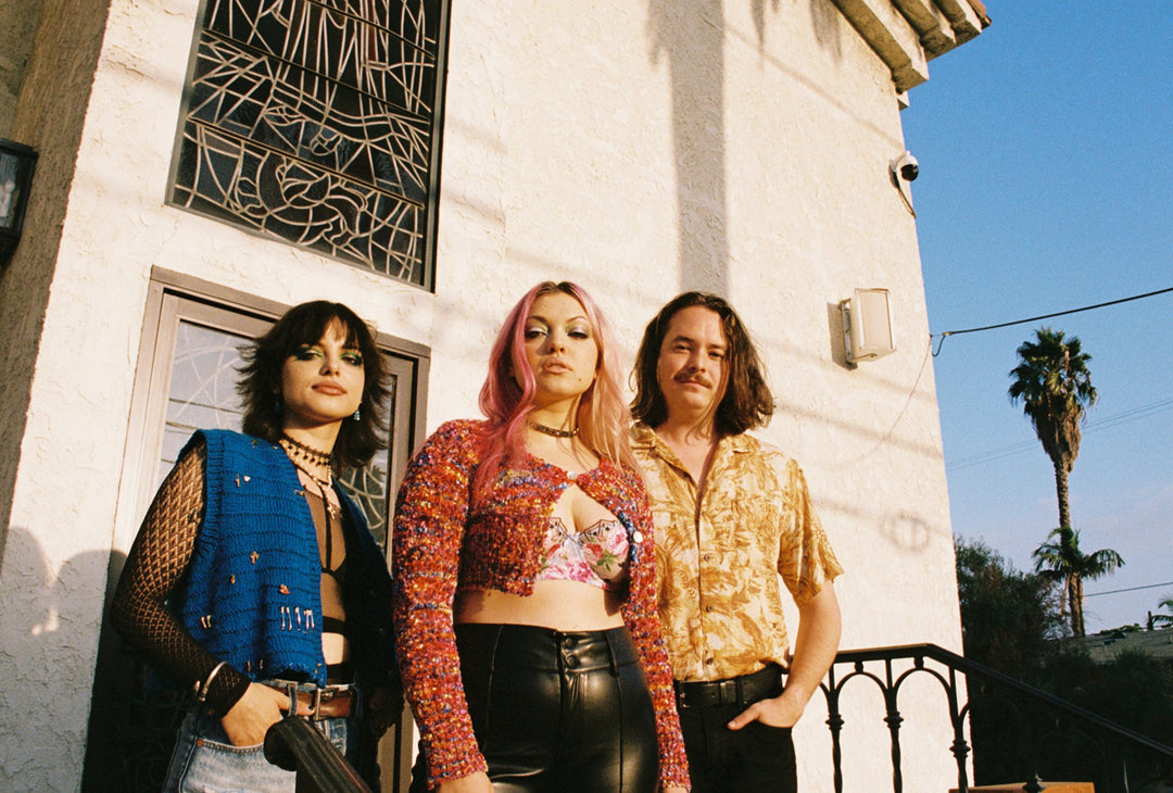 Three people stand outside a light-colored building with stained glass and a palm tree nearby. They wear colorful, retro-inspired clothes and look confidently at the camera in the afternoon sun.