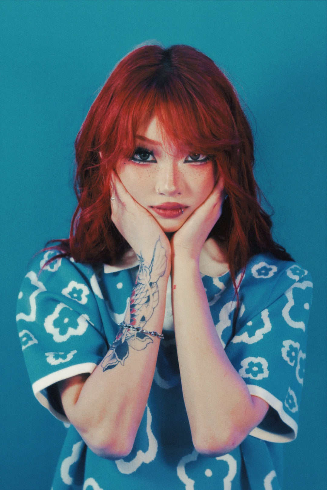 A person with red hair poses against a blue background, wearing a blue shirt with white flower patterns and resting their face in their hands. A tattoo is visible on their forearm.