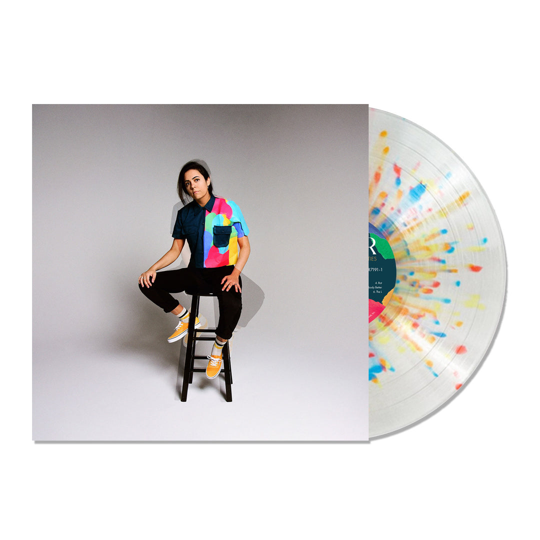 A Limited Edition Illuminati Hotties vinyl with a colorful splatter design partially slides out of a sleeve. The sleeve shows a person on a stool in a vibrant multicolored shirt and yellow sneakers against a plain background.