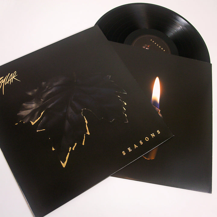 A vinyl record by Sylar is partially pulled from its sleeve, which has a black cover with a dark leaf and SEASONS in gold. The inner sleeve shows a flame on black, hinting at the evocative tracks inside this music album.
