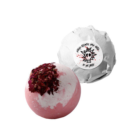 A pink and white Stand Atlantic bath bomb with shea butter and dried rose petals sits beside its white paper packaging, featuring a punk-style graphic and text label.