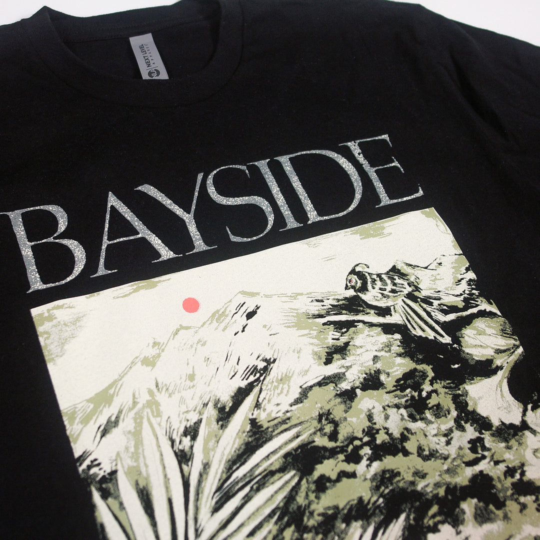 A black Bayside t-shirt displays BAYSIDE in bold white letters with a bird on rocks, mountains, plants, and a red sun—a unique design available only here.