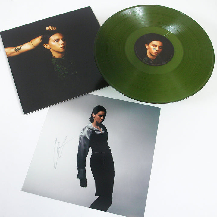 A green PVRIS vinyl record, its signed 12x12 cover featuring a short-haired person with an arm raised, and a separate photo of them in black and white attire are displayed on a white surface.