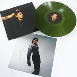 A green PVRIS vinyl record, its signed 12x12 cover featuring a short-haired person with an arm raised, and a separate photo of them in black and white attire are displayed on a white surface.