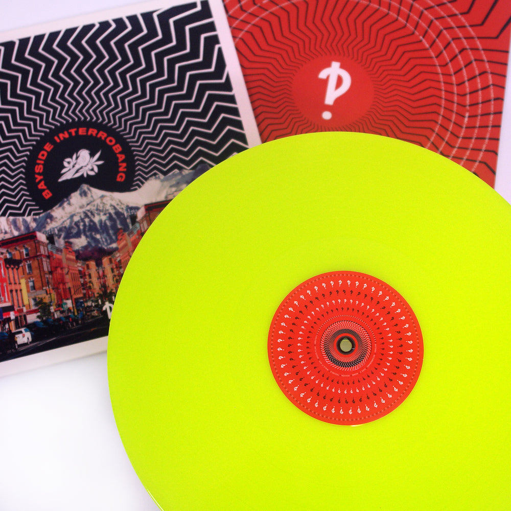 A Neon Yellow Bayside LP with a red and black patterned label sits in front of two album covers, one showcasing mountain and city scenes, the other featuring a red geometric design from their Interrobang release.
