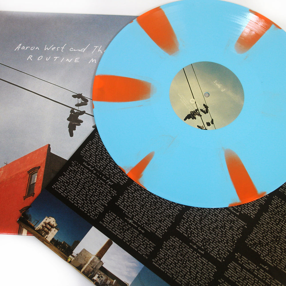 A blue vinyl with orange splashes rests on an album cover featuring Aaron West and the Roaring Twenties’ birds perched on power lines by a red building, with a black inner sleeve showing white Pinwheel Tracklisting text.