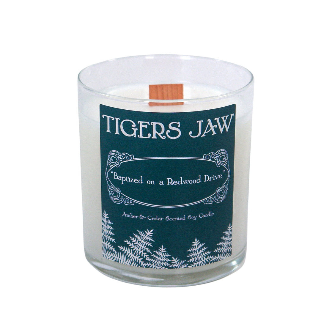 A white Tigers Jaw candle in a clear jar labeled "Baptized on a Redwood Drive" features soothing amber and cedar scents, with fern illustrations along the bottom of the label.