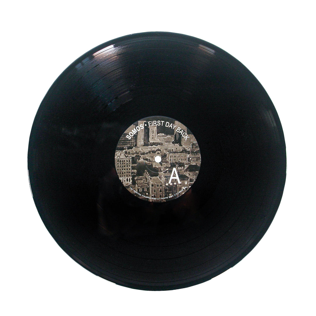 A black vinyl record by Somos with a center label showing a sepia cityscape, the text First Day Back, a large letter A, and a subtle Black Backpack motif on the right side.