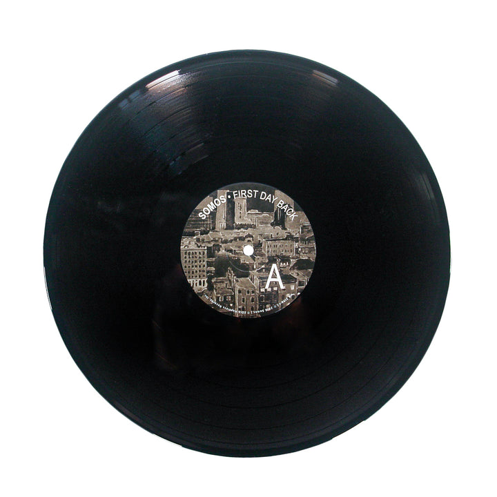 A black vinyl record by Somos with a center label showing a sepia cityscape, the text First Day Back, a large letter A, and a subtle Black Backpack motif on the right side.