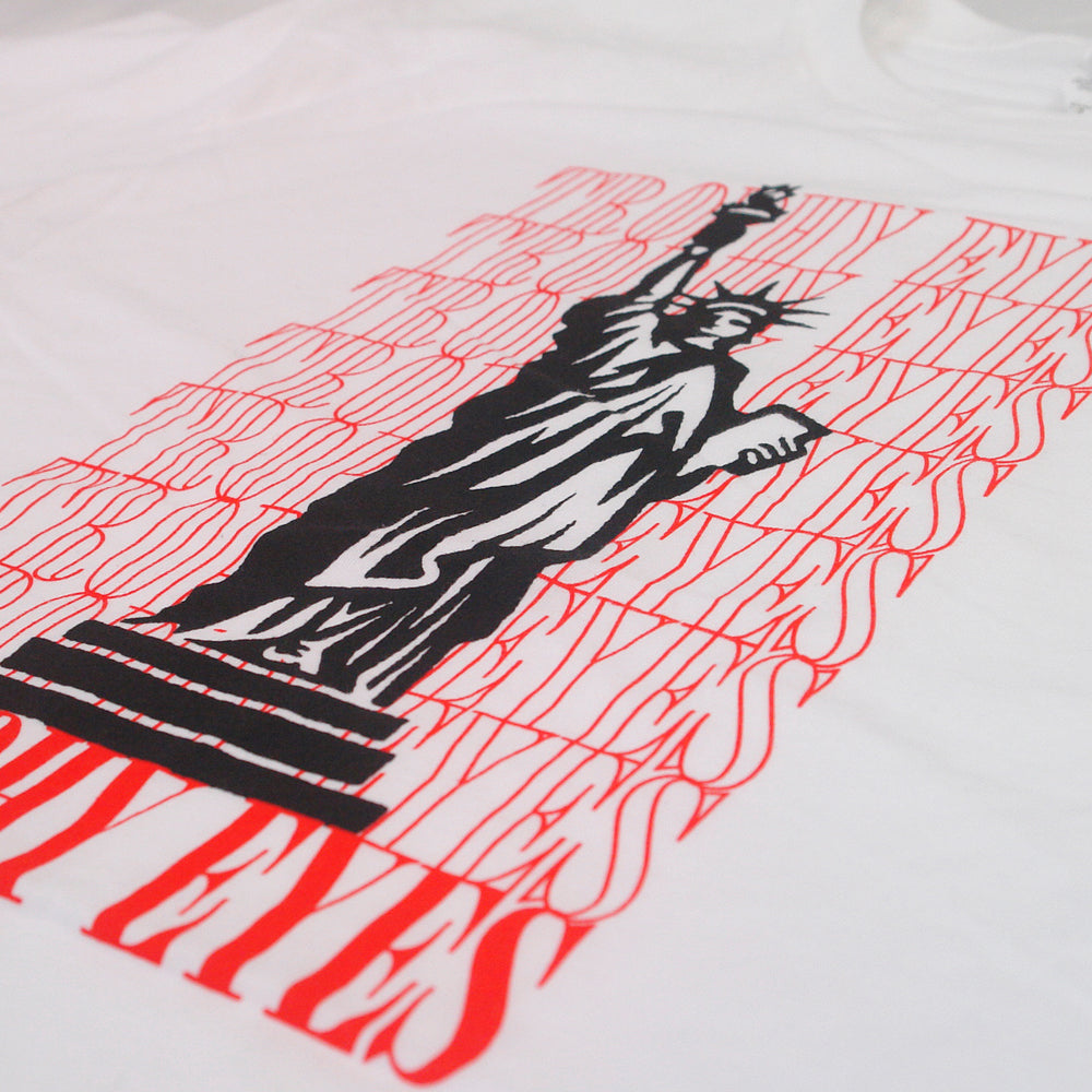 A Trophy Eyes White T-shirt showcases a black graphic of the Statue of Liberty, overlaid with repeated red DRY EYES text in a vertical, distorted font—this unique design makes a bold statement.