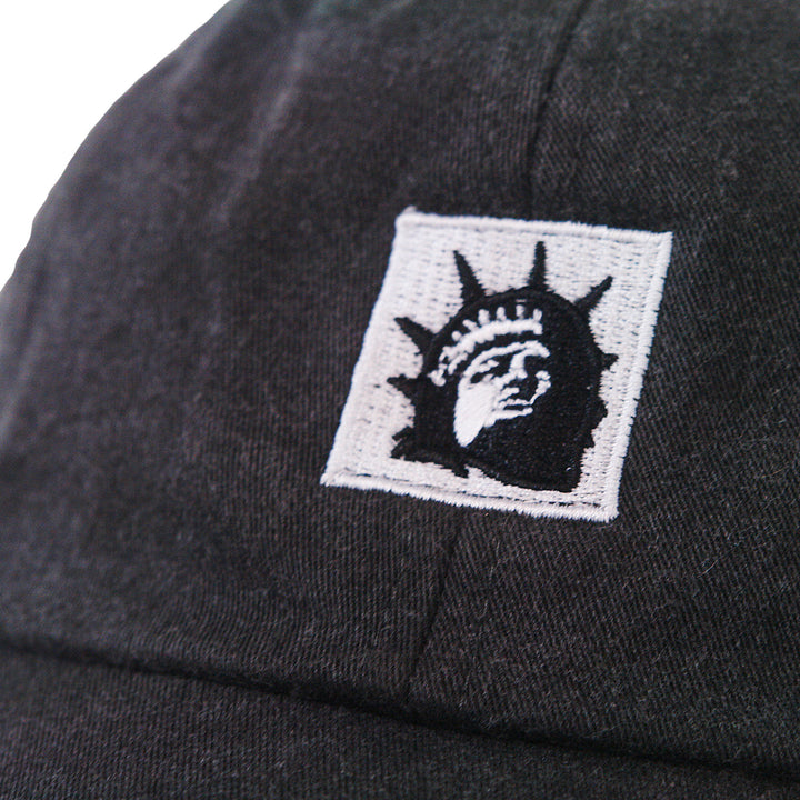 Close-up of a black Trophy Eyes Dad Hat with an embroidered patch showing a stylized black and white Statue of Liberty face.