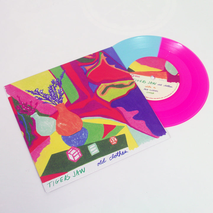 A blue and neon magenta vinyl record by Tigers Jaw partially slides out of a colorful album cover with abstract shapes, vases, dice, and handwritten tracklisting. The cover reads old clothes in handwritten text.