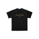 Exclusive Between You And Me design on a black Next Level tee: WELCOME TO THE ARMAGEDDON in yellow, centered above a small purple atom graphic. Plain white background.