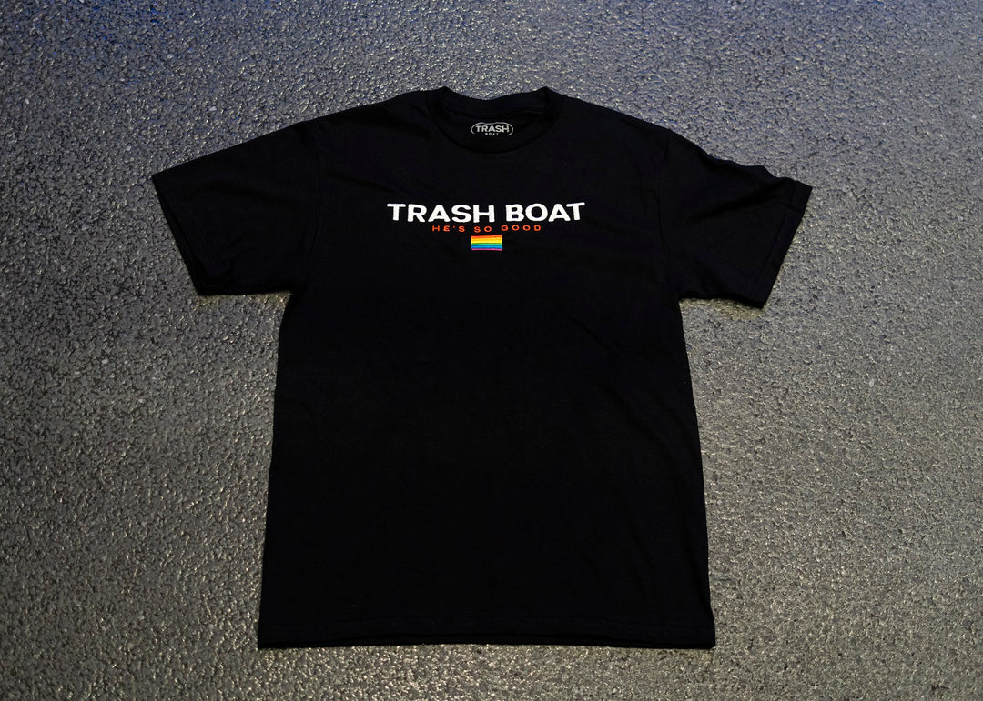 A black Trash Boat shirt is laid flat on a gray surface with TRASH BOAT in large white letters, HE’S SO GOOD in smaller orange text, and a colored bar underneath. Available in multiple sizes, including X-Large. Brand: Trash Boat.