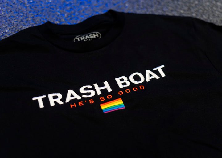 Trash Boat black shirt with bold white “TRASH BOAT,” red “HES SO GOOD,” and a small rainbow flag, shown on a textured blue background. Available in sizes up to X-Large.
