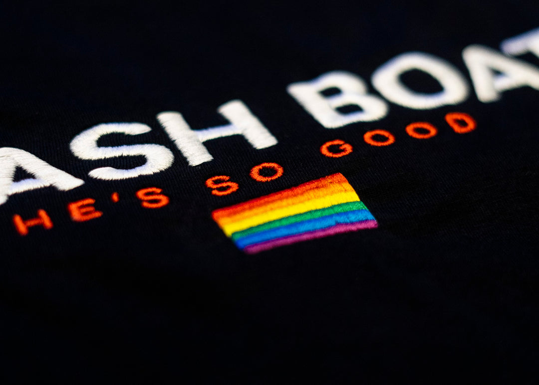 Close-up of white and orange ASH BOA and HES SO GOOD text embroidery with a rainbow flag below on a Trash Boat shirt. Available in X-Large and other sizes. Brand: Trash Boat.
