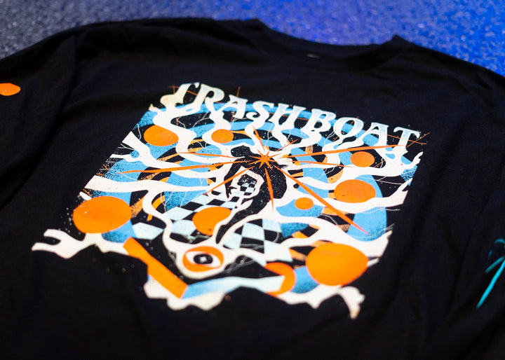 A black Trash Boat merch T-shirt with an abstract, colorful design in orange, blue, and white shapes centered around TRASH BOAT in bold white letters. Available as a tee or long sleeve.