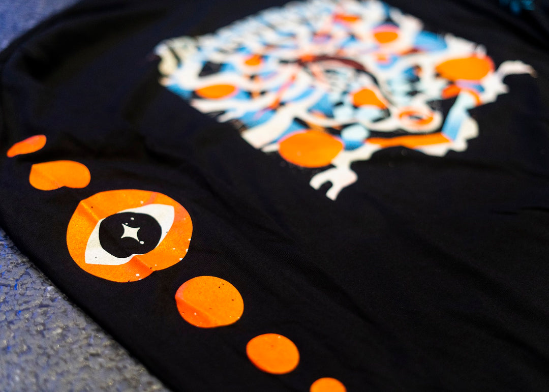 A black Trash Boat long sleeve with a vibrant abstract design in white, blue, orange, and red at the center, plus orange circles and stylized eye graphics along the side—an ideal addition to any band merch collection.