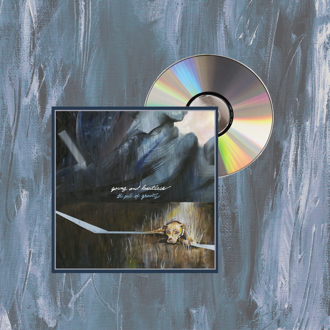 A CD by Young And Heartless, partly in its case, rests on a blue and gray painted background. Its cover shows abstract brushstrokes, a yellow dog’s face below, and white handwritten text at the center referencing Pull Of Gravity tracks.
