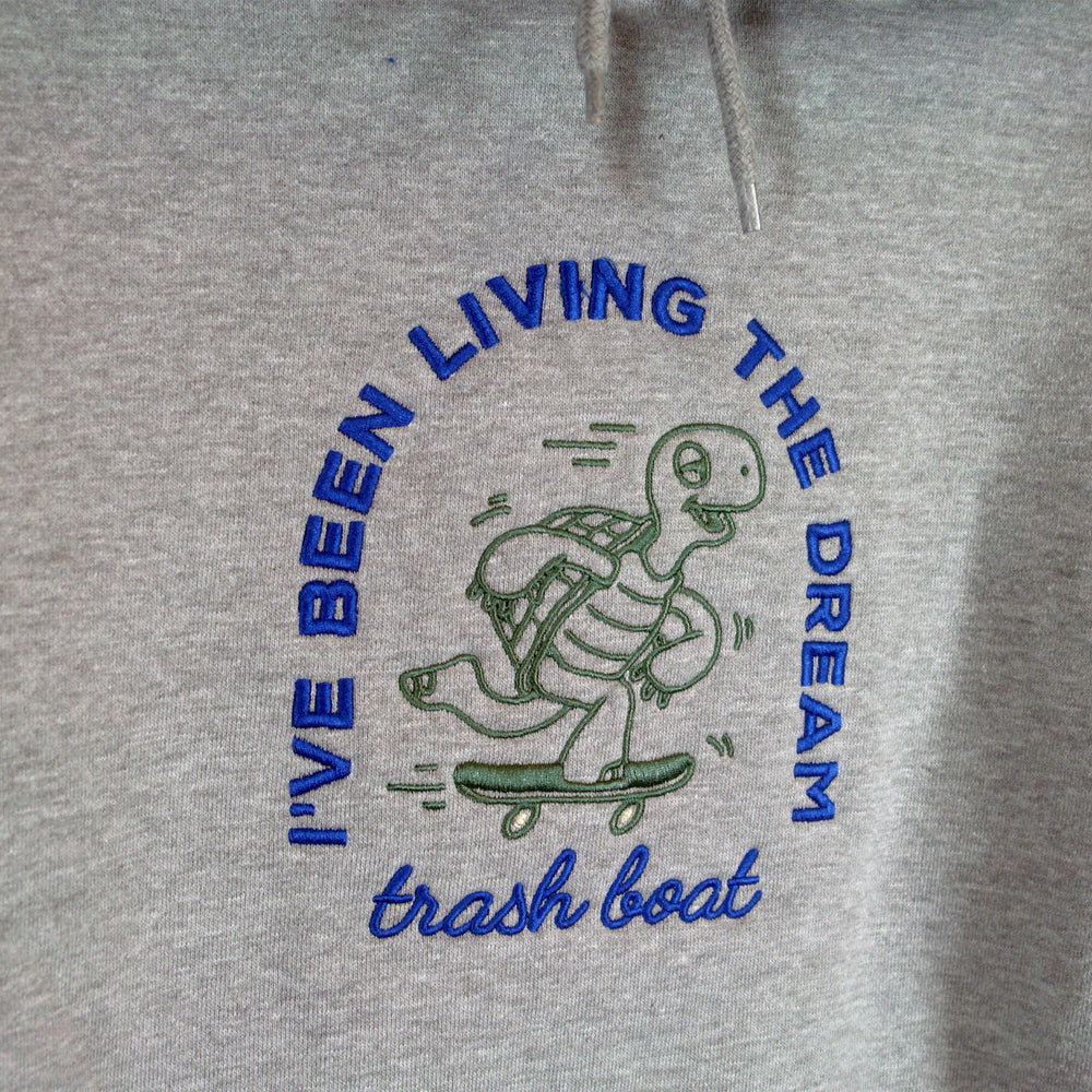 Gray hoodie featuring a green cartoon turtle skateboarding with blue “I’VE BEEN LIVING THE DREAM” text and cursive trash boat below. Brand: Trash Boat.