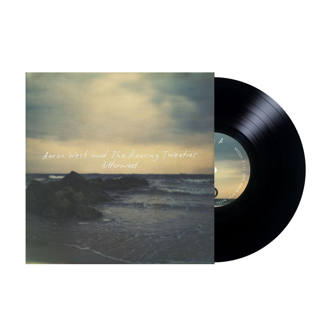 A limited edition vinyl partially slides from its cover, which features a blurry seashore with rocks and water. White handwritten-style text reads “Aaron West and the Roaring Twenties Bittersweet Black.”.