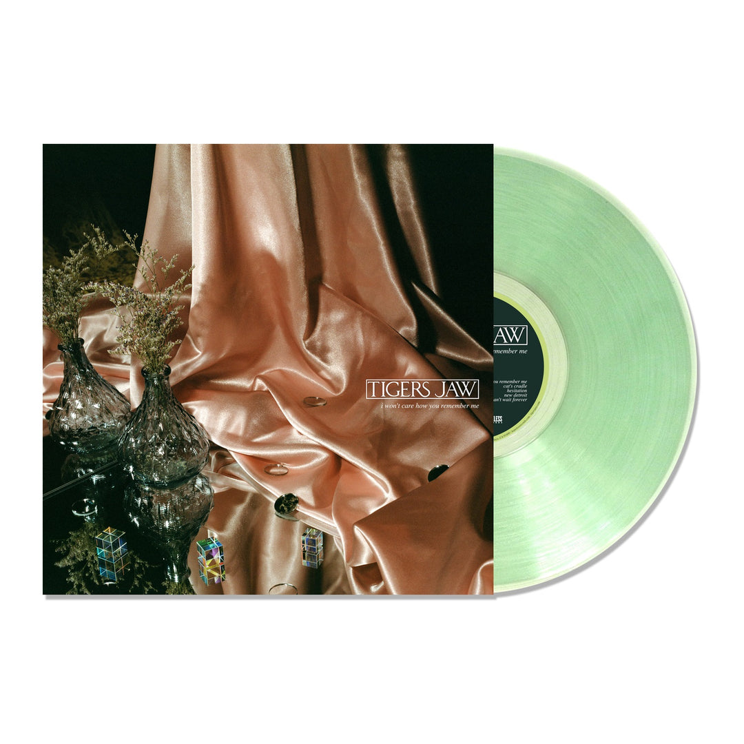 A limited edition green vinyl record by Tigers Jaw is partially pulled from its album cover, which displays metallic pink fabric, glass vases, and decorative objects arranged on a reflective surface.
